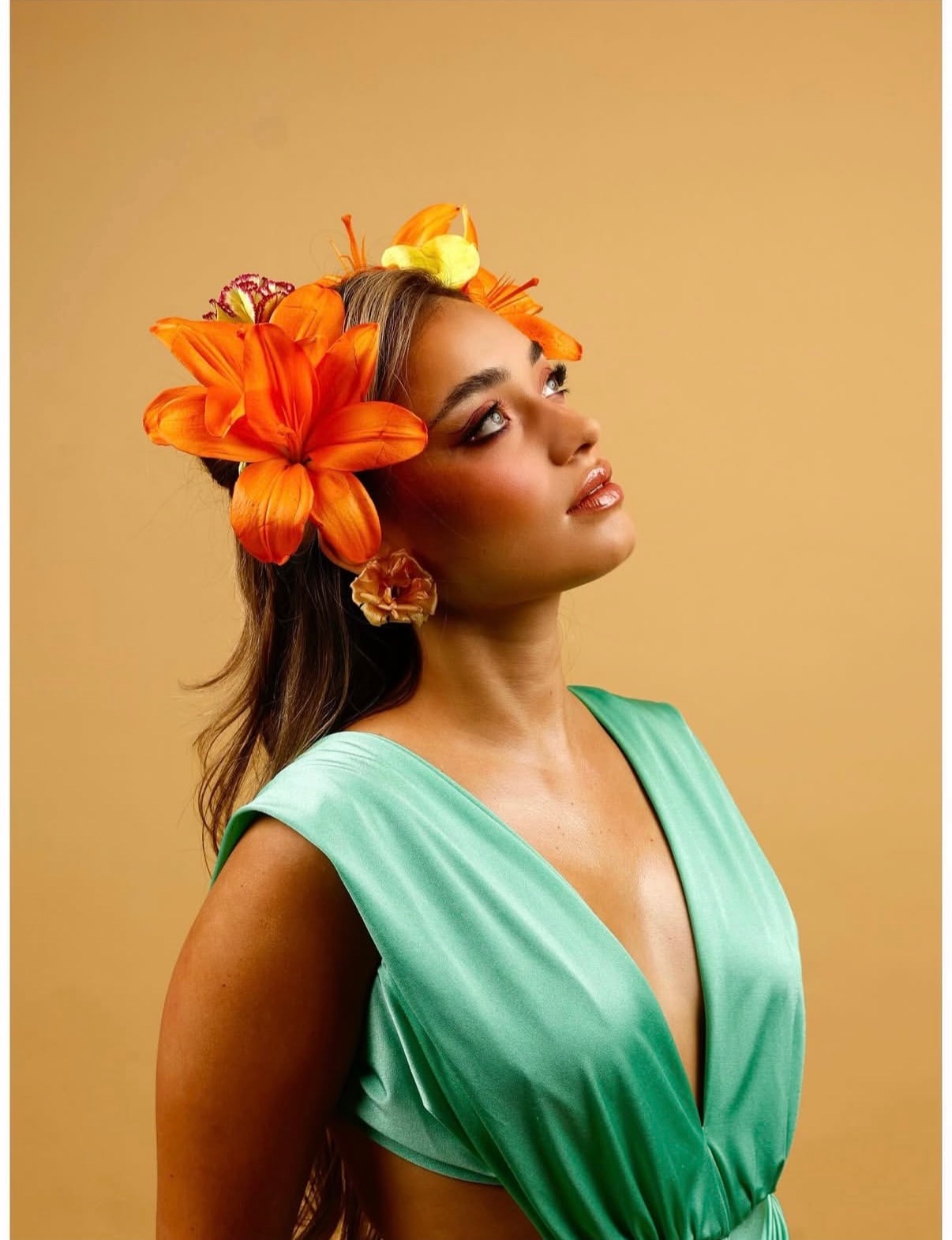 Beauty editorial portrait: a model in a green dress with bright orange lilies in her hair, lit against a warm ochre backdrop.