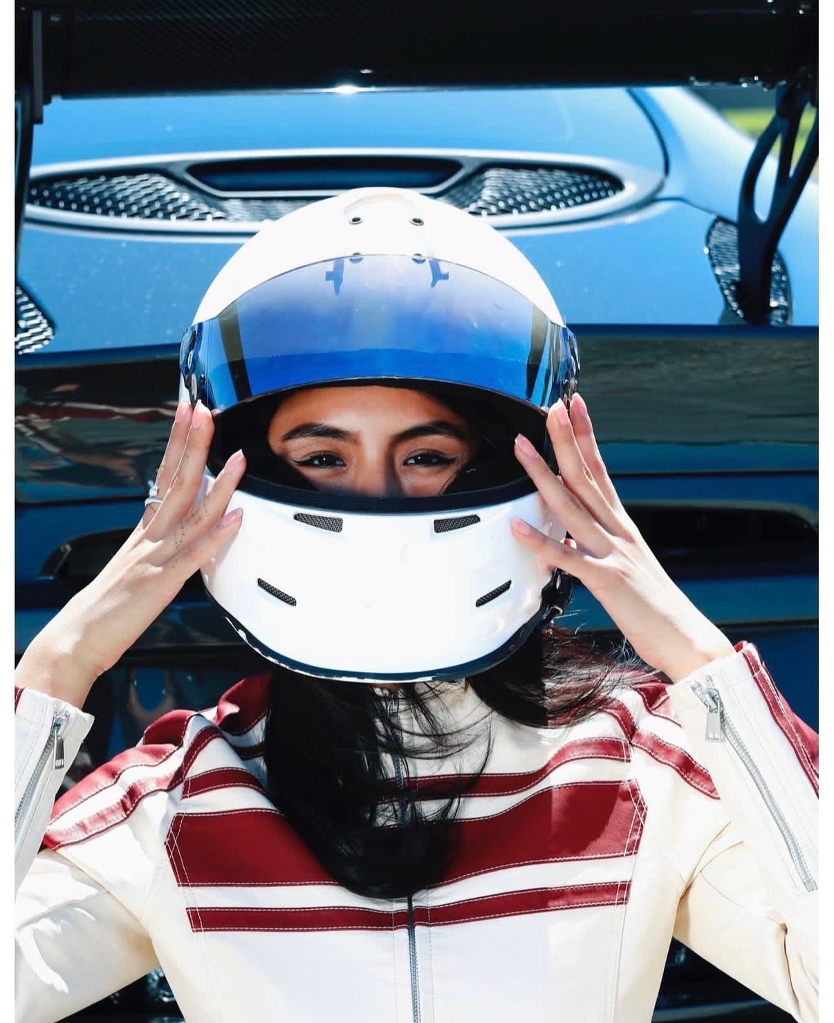 Automotive lifestyle portrait: a driver in a cream and red racing suit lowering a white open-face helmet, a dark sports car behind her.