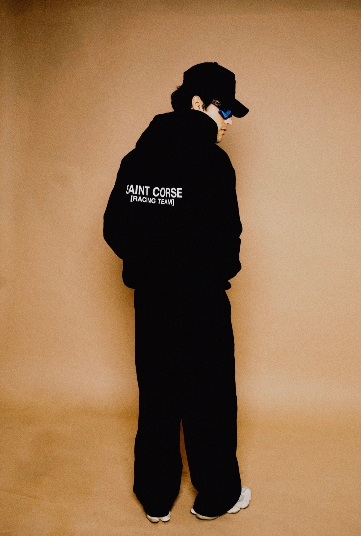 Brand apparel photograph for Saint Corse Racing Team: a figure in an all-black tracksuit and cap turned away from camera against a tan seamless backdrop.